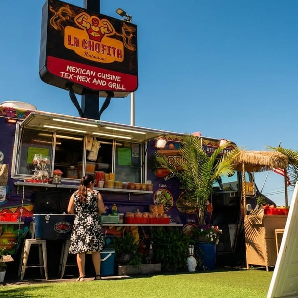 A woman stands at the counter of a colorful La Chofita Mexican food truck on a sunny day.