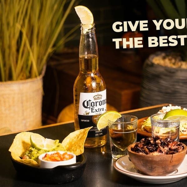 Corona beer, tequila shots, guacamole, chips, salsa, and tacos on a dark table.