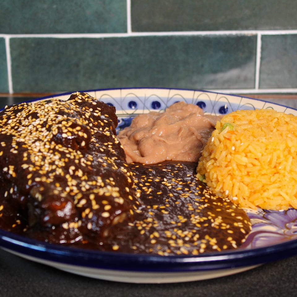 Mole-covered chicken topped with sesame seeds, orange rice, and refried beans on a decorative plate.