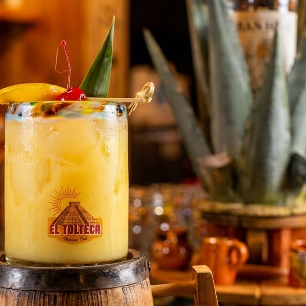 A vibrant yellow cocktail with a cherry and pineapple garnish, branded "El Tolteca Mexican Food".