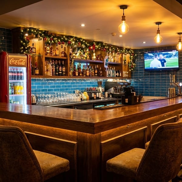 A cozy bar with a wooden counter, bar stools, a Coca-Cola fridge, and festive garland.