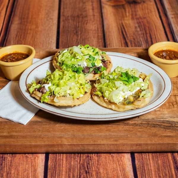 Three sopes topped with lettuce, cheese, and meat, served with two small bowls of salsa on a wooden tray.