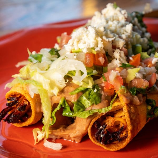 Three golden taquitos topped with lettuce, pico de gallo, refried beans, and crumbled white cheese.