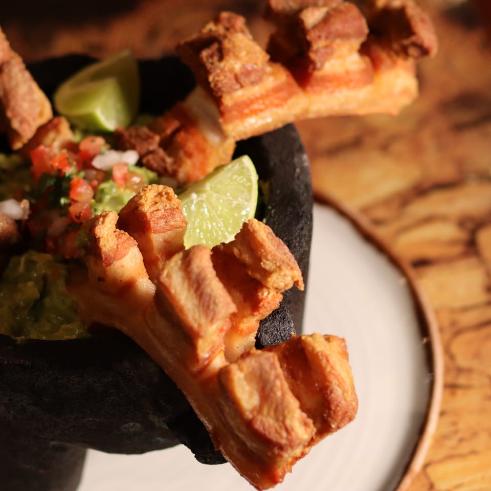 Crispy pork belly pieces with guacamole, pico de gallo, and lime in a black serving dish beside a wooden board.