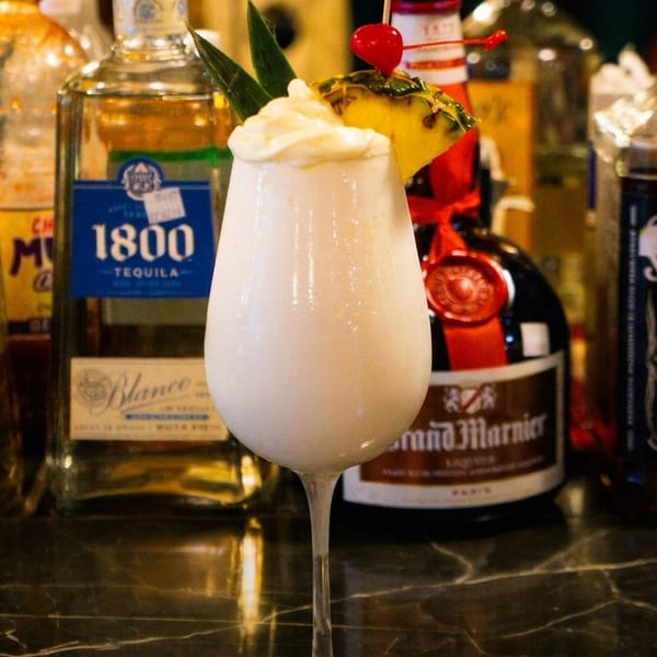 A creamy white cocktail with pineapple and a cherry garnish, surrounded by liquor bottles on a bar.