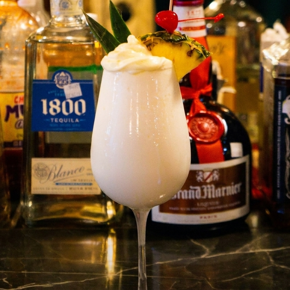 A creamy white cocktail with pineapple and a cherry garnish, surrounded by liquor bottles on a bar.