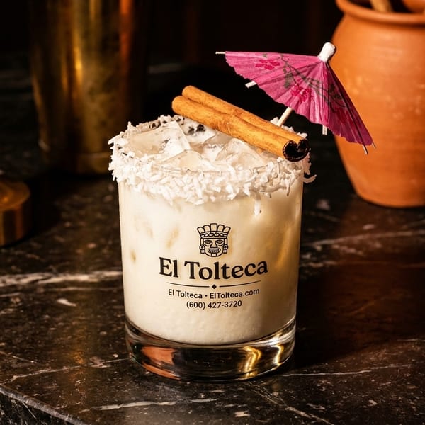 El Tolteca cocktail with coconut rim, cinnamon sticks, and a pink umbrella on a marble bar.